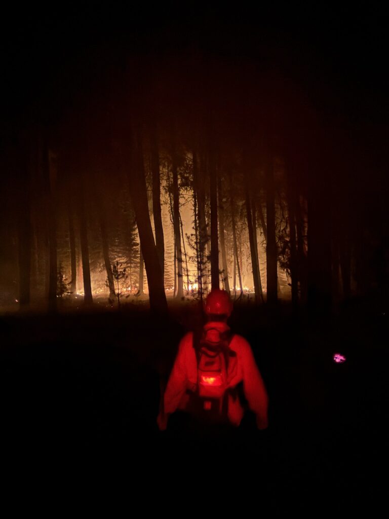 A firefighter in the foreground with flames from a fire in the background. It is night time