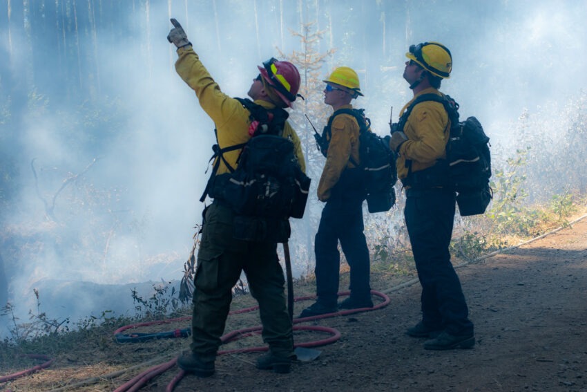 Three firefighters standing. One is pointing up toward the sky.