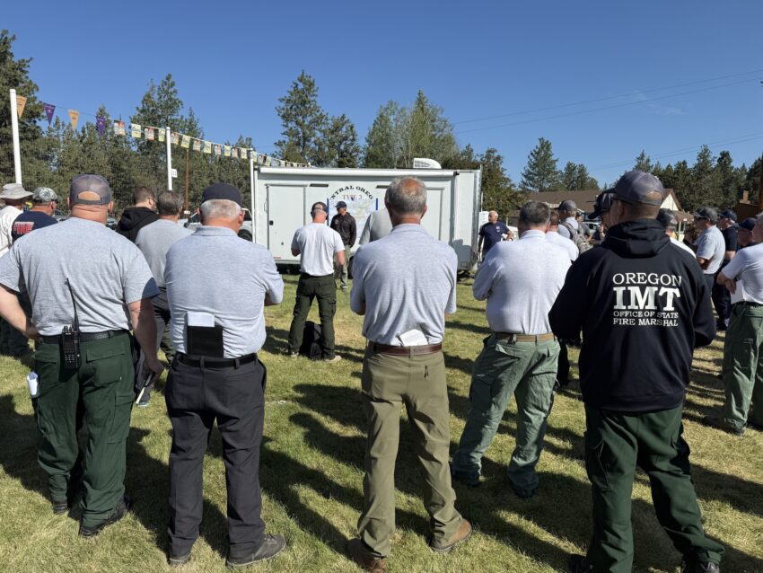 Many firefighters stand facing a person speaking by a trailer at a fire briefing on the Alder Springs Fire.