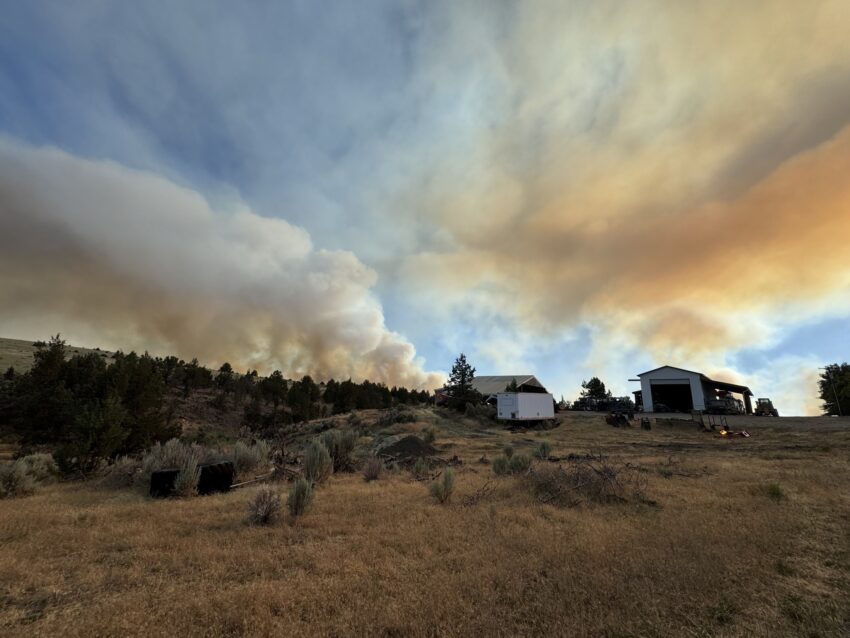 Photo of a landscape with a blue sky and heavy smoke cutting through it. A home and another building sit in the distance.
