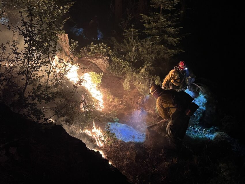 Two firefighters work to dig line at the Elk Fire in the dark. The fire is blazing brightly.