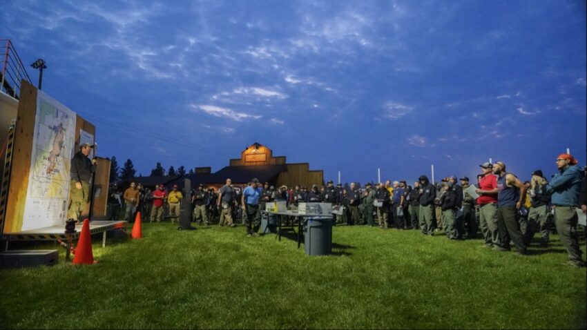 Fire briefing at the Flat Fire. A man stands near fire maps and speaks into a microphone at a crowd of firefighters looking on.