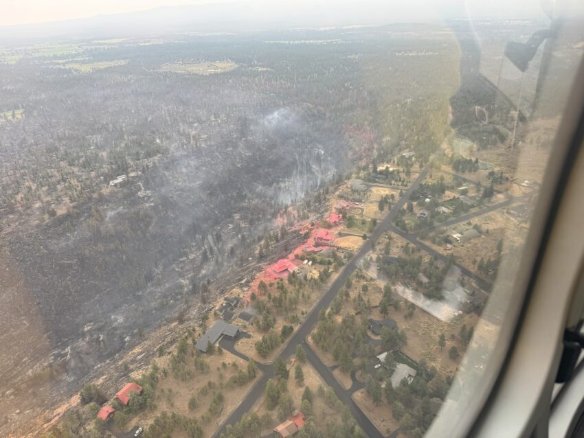 Aerial photo of the Flat Fire in Central Oregon. There are homes on a ridge with burnt land to one side and a line of fire retardant between them.