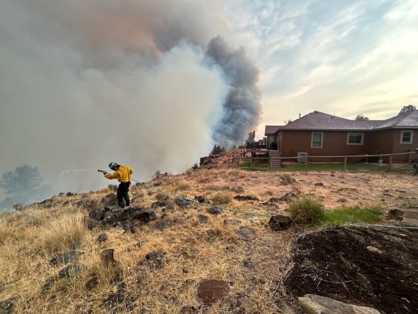 Firefighter shoots a water hose at the Flat Fire with a plume of smoke and a house in the background.